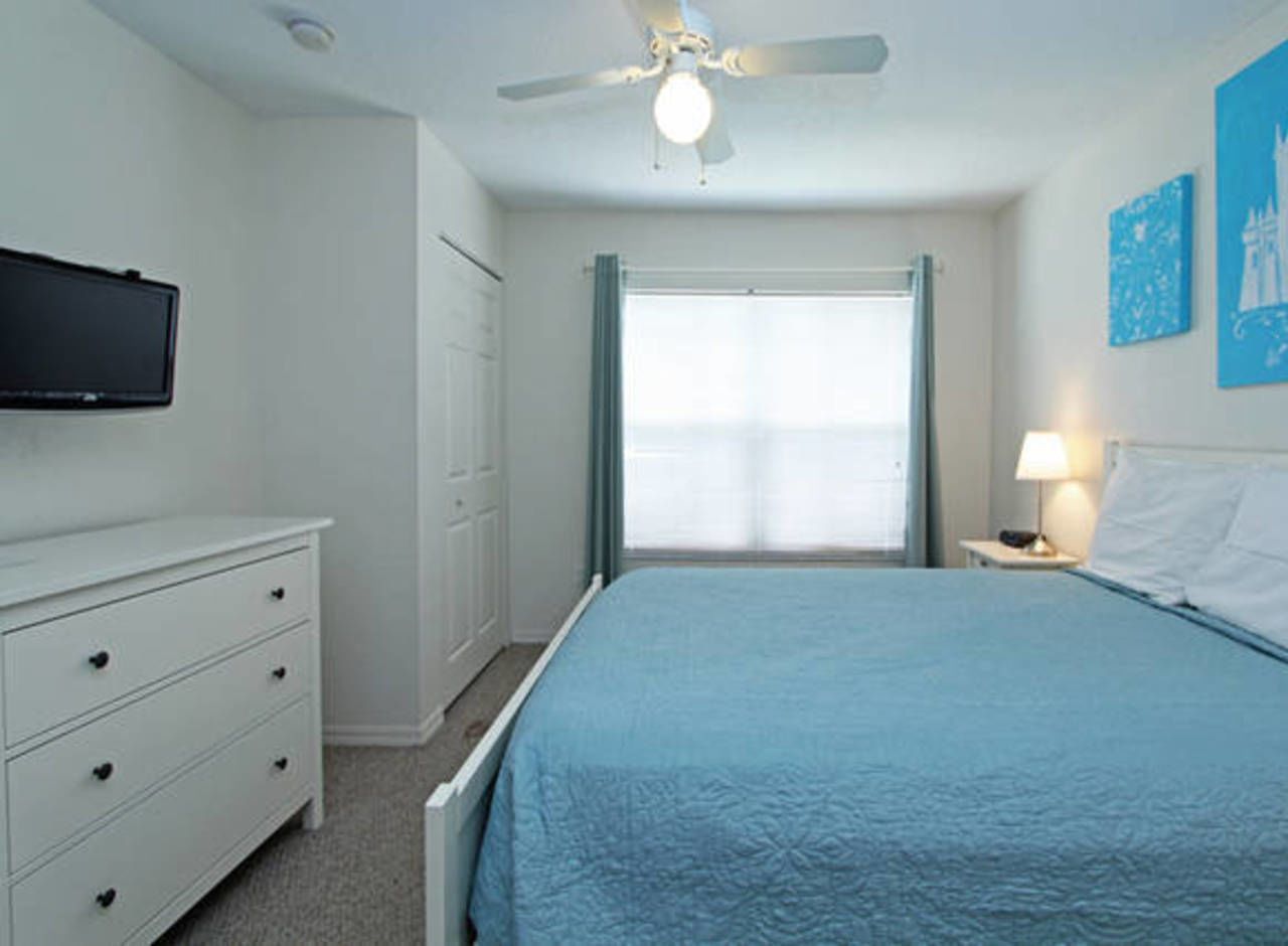 A bedroom with a bed, a dresser, a ceiling fan, a window with curtains, and a wall-mounted TV.