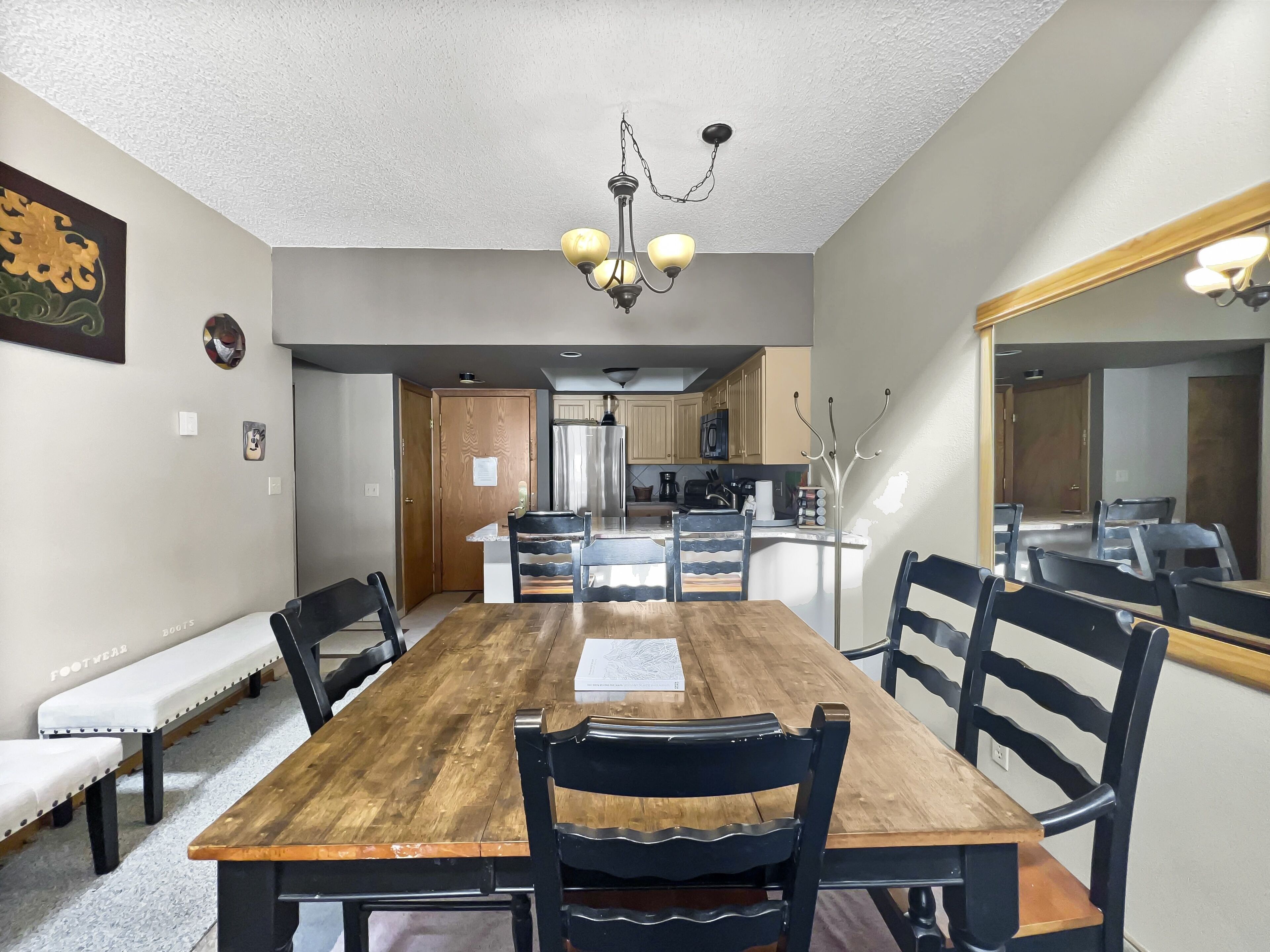 A dining area with a wooden table and chairs, a chandelier, and a kitchen in the background.