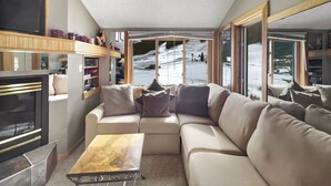 A cozy living room with a sectional sofa, a fireplace, and a view of a snowy landscape through large windows.
