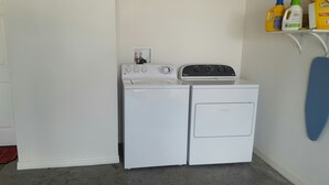 A laundry room with a washer and dryer, a shelf with cleaning supplies, and a blue towel hanging on the wall. - Patio Villa in Duval Village - Gas Golf Cart INCLUDED - 5 minutes to everywhere (The Villages)