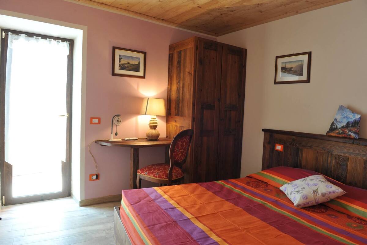 A bedroom with a bed, a chair, a desk with a lamp, and wooden wardrobe.