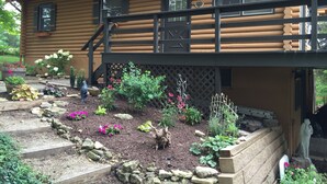 Una cabaña de troncos con terraza, rodeada por un jardín con diversas plantas y flores.