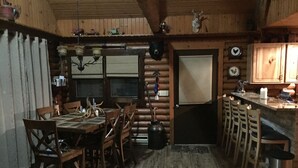 Interior rústico de una cabaña de madera con comedor, cocina y un amplio ventanal.