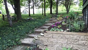 Un jardín con escalones de piedra que conducen a una terraza de madera, rodeado de árboles y diversas plantas.