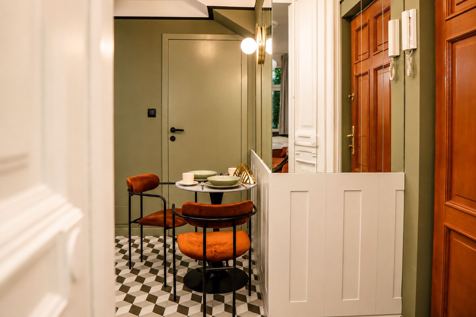 A narrow hallway with a checkered floor leading to a dining area with two chairs and a table set with plates, cups, and a candle.