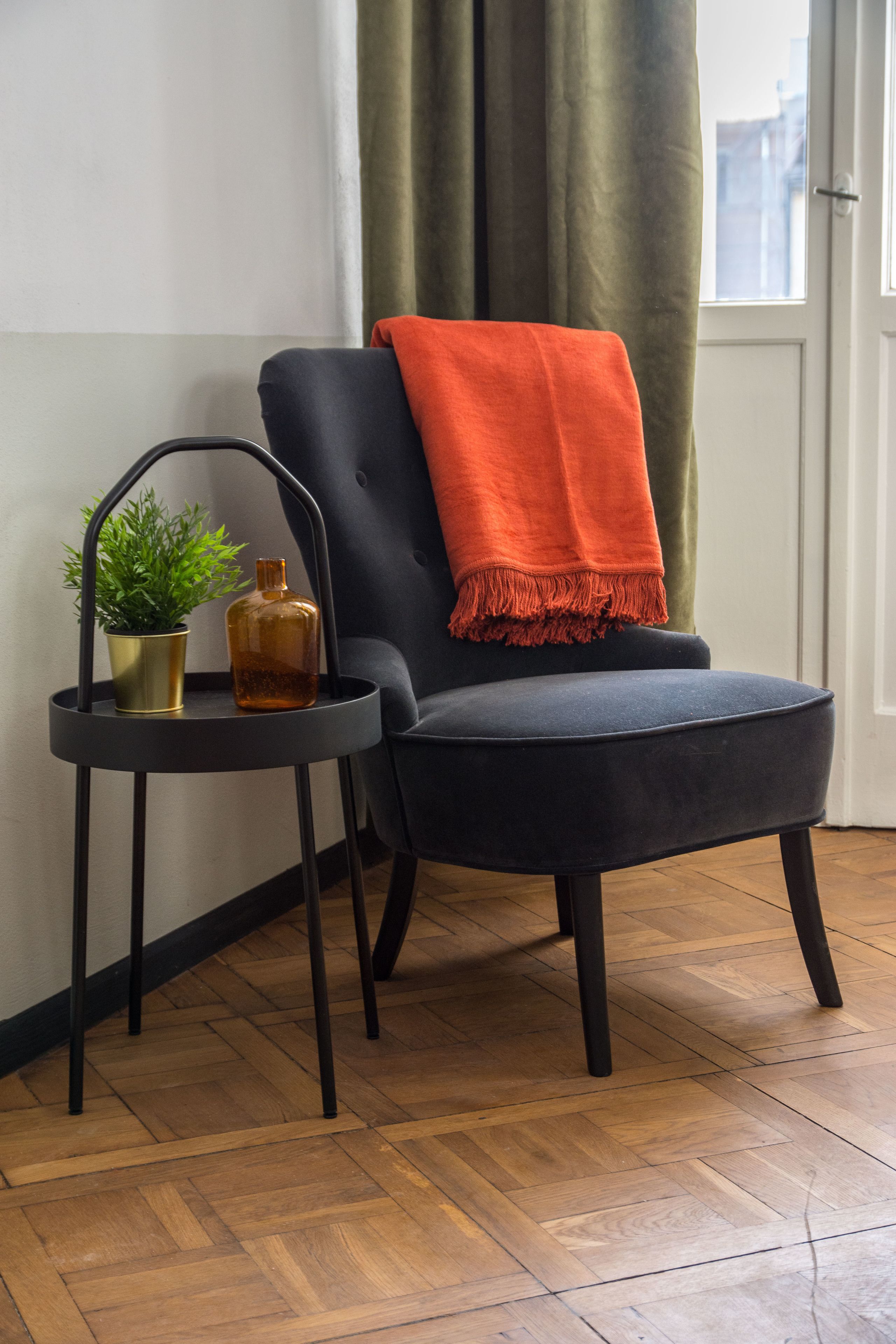 A chair with a dark fabric and an orange throw blanket, a small table with a potted plant and a bottle, and a wooden parquet floor.