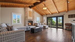 A spacious living room with a fireplace, wooden ceiling beams, and large windows.
