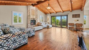 A spacious living room with a fireplace, wooden ceiling beams, and a large sliding glass door leading to a balcony.