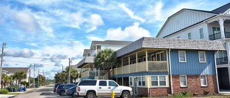 A street view with a two-story building featuring a balcony, parked cars, and a clear sky.