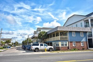 A street view with a two-story building featuring a balcony, parked cars, and a clear sky. - 202 28th Ave Unit 6 1 Bedroom Condo (North Myrtle Beach)