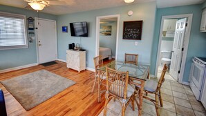 A spacious living room with a glass dining table, wicker chairs, a ceiling fan, and a television mounted on the wall. - 202 28th Ave Unit 6 1 Bedroom Condo (North Myrtle Beach)