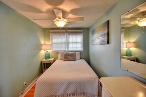 A bedroom with a bed, a ceiling fan, two bedside tables with lamps, a window with blinds, and a painting on the wall. - 202 28th Ave Unit 6 1 Bedroom Condo (North Myrtle Beach)