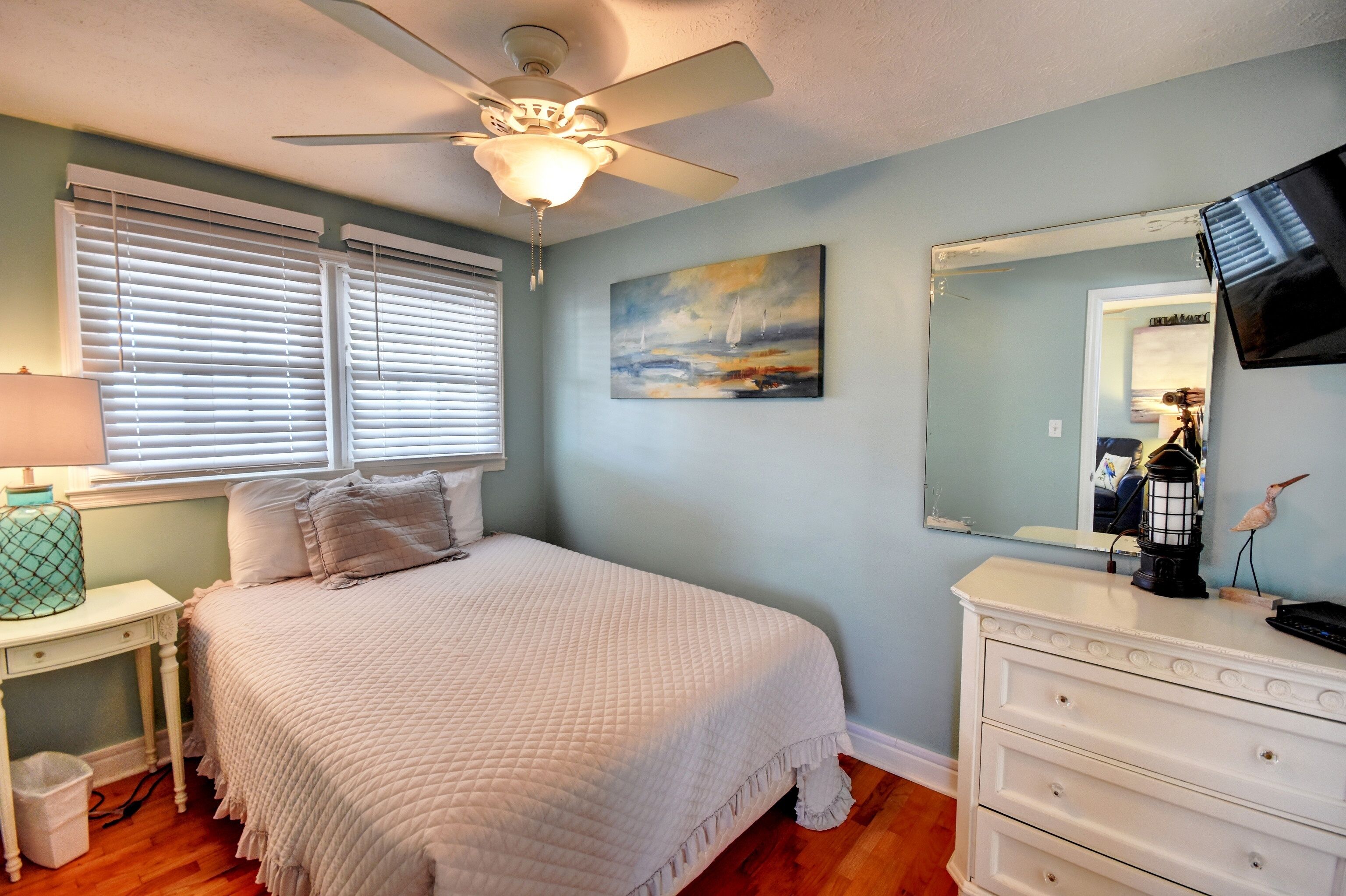 A bedroom with a bed, a nightstand, a dresser, a ceiling fan, a painting, and a mirror.