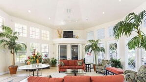 A spacious living room with a large orange sofa, a coffee table, and several chairs. There are multiple potted plants and a large window providing natural light. - Silver Palm Retreat (Kissimmee)