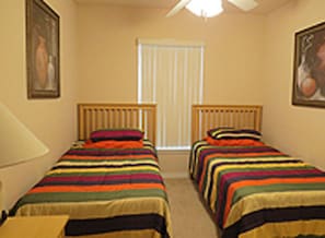 Quarto com duas camas de solteiro, cada uma com colcha listrada e cabeceira de madeira. Há uma janela com persianas, um ventilador de teto e um quadro na parede.