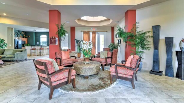 A spacious lobby with a central seating area featuring a round coffee table surrounded by four chairs. The lobby has a high ceiling with a skylight and is decorated with potted plants and artwork. - Ridge Castle (Davenport)