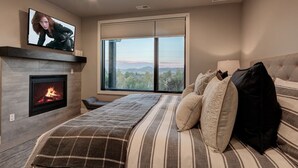 A bedroom with a fireplace, a large window with a view, a bed with striped bedding, and a television on the wall.