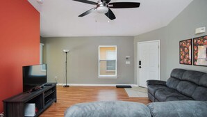 Ein Wohnzimmer mit Deckenventilator, Fernseher auf Holzfuß, grauem Sofa und Stehlampe.