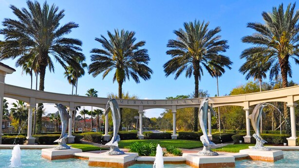 A fountain with sculptures, palm trees, and a pergola with the text "MERALD ISLAND". - Jewel in the Crown (Kissimmee)