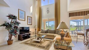 A spacious living room with a large TV, comfortable seating, a potted plant, and a view of the outdoors. - Jewel in the Crown (Kissimmee)