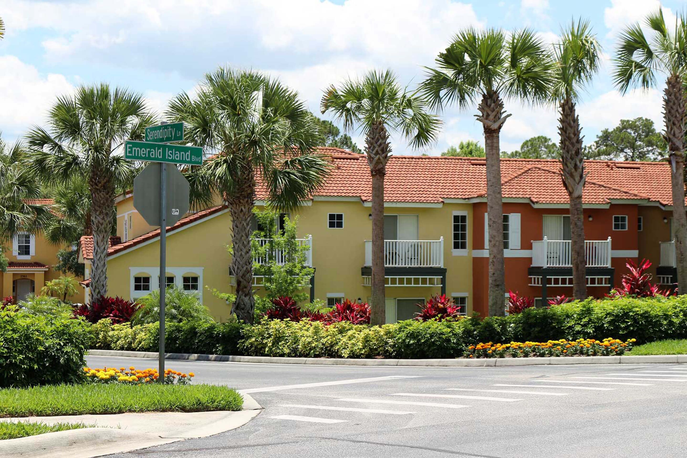A yellow building with balconies, palm trees, and a stop sign at the intersection of Emerald Island Blvd and Serendipity Blvd.