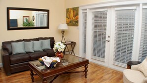 A living room with a brown leather sofa, a glass coffee table, and a beige armchair. There is a painting on the wall and a door with blinds. - Heritage Haven (Kissimmee)
