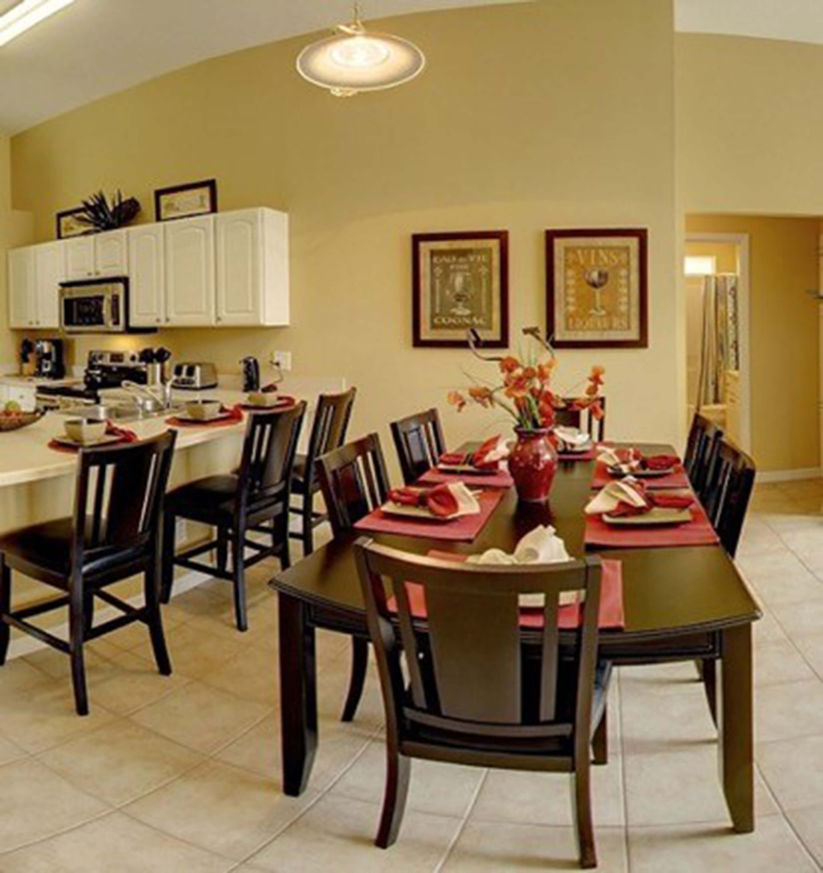 A dining area with a dark wooden table set for six, surrounded by chairs. There is a kitchen area with white cabinets and a microwave visible in the background.
