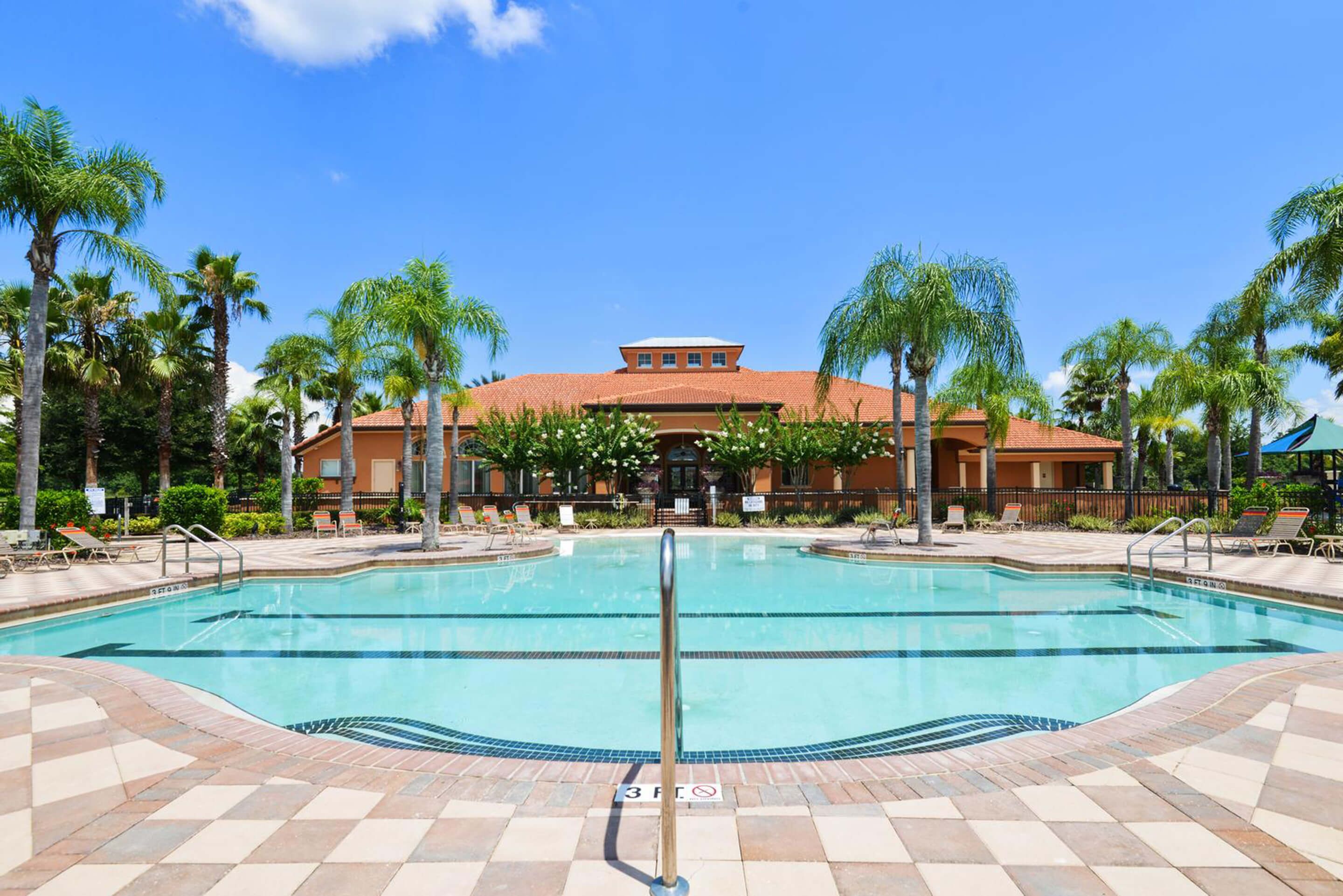A swimming pool with a tiled deck, surrounded by palm trees and a building with a red roof in the background.