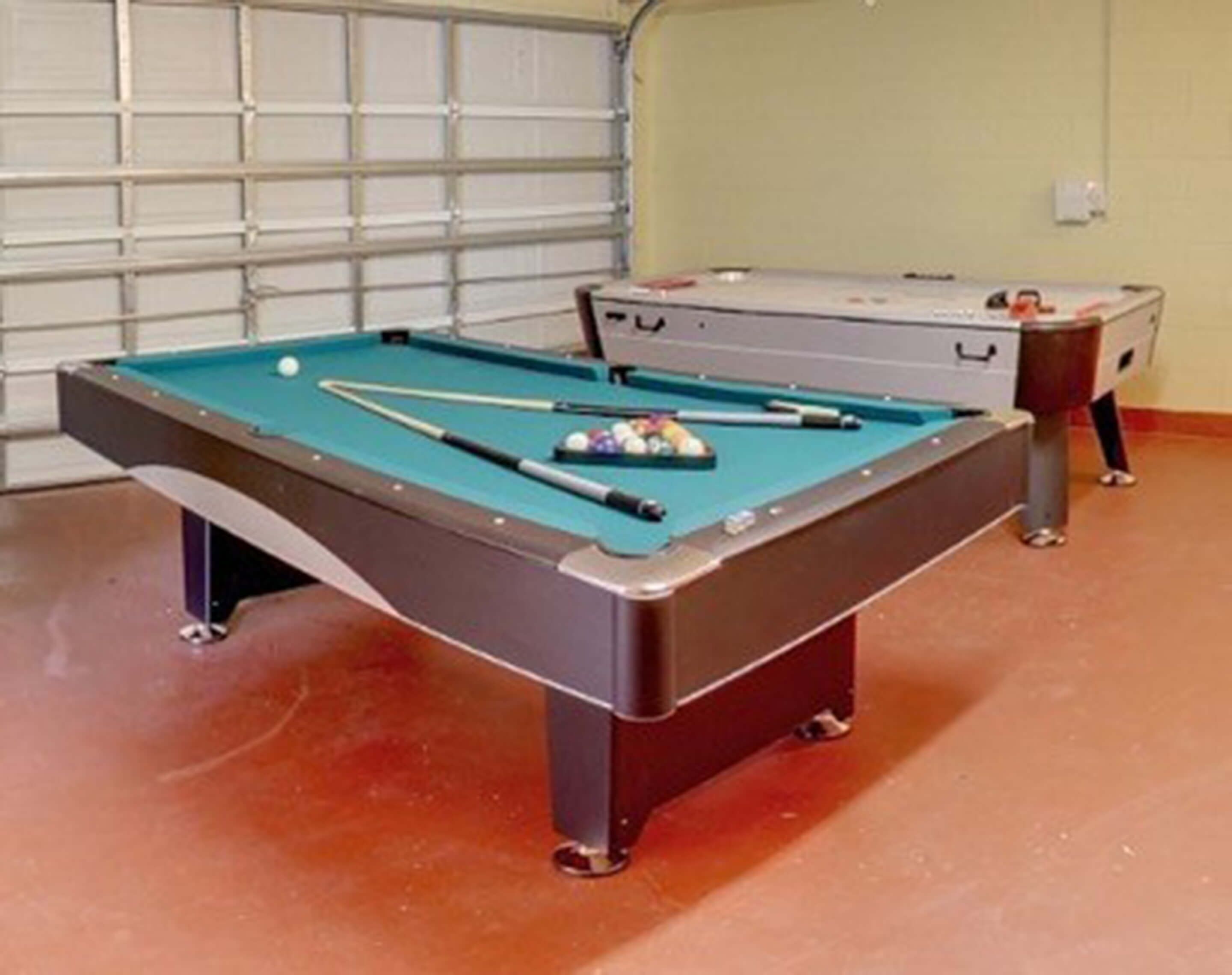 Ein Billardtisch mit Kugeln und Spielstöcken, ein Koffer und im Hintergrund eine Garagentür.