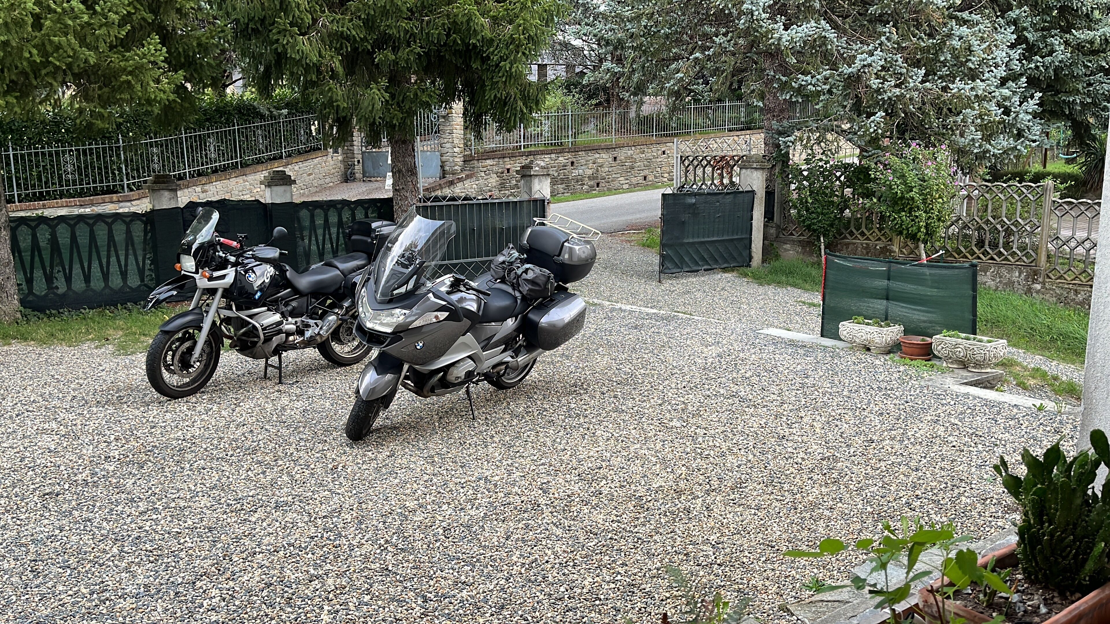 Deux motos sont garées sur une allée de gravier, avec un mur de pierres et une clôture métallique en arrière-plan.