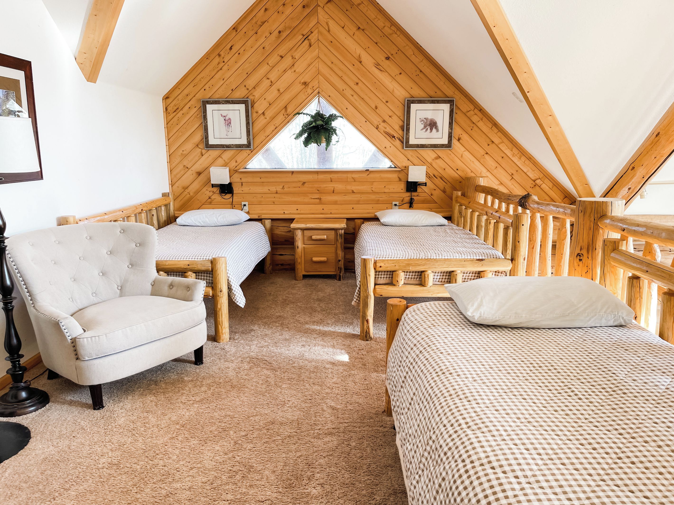 A cozy room with two beds, a chair, and a desk, featuring wooden walls and a sloped ceiling.