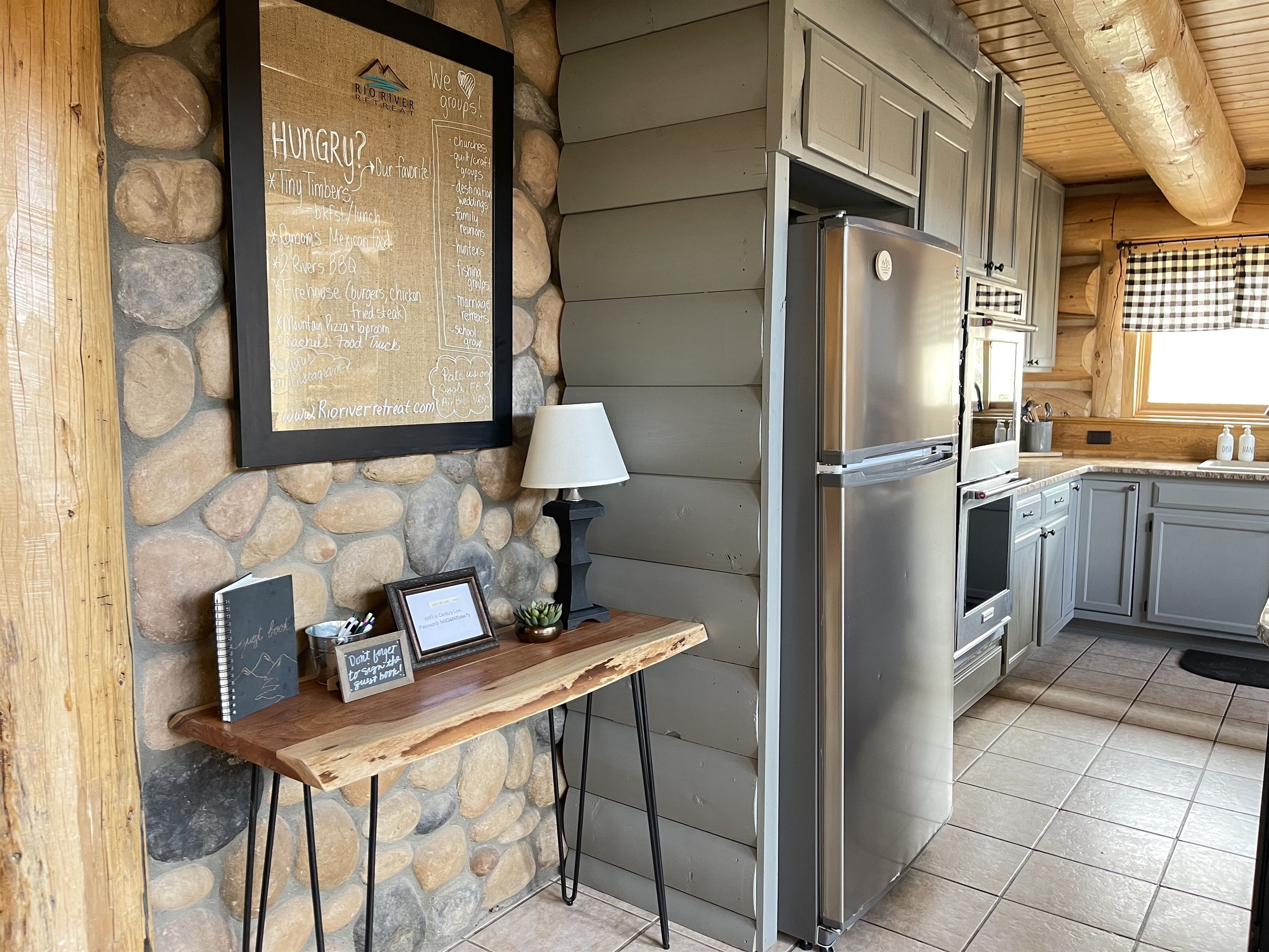 A kitchen with a stone wall, a framed menu, a laptop, and a refrigerator.