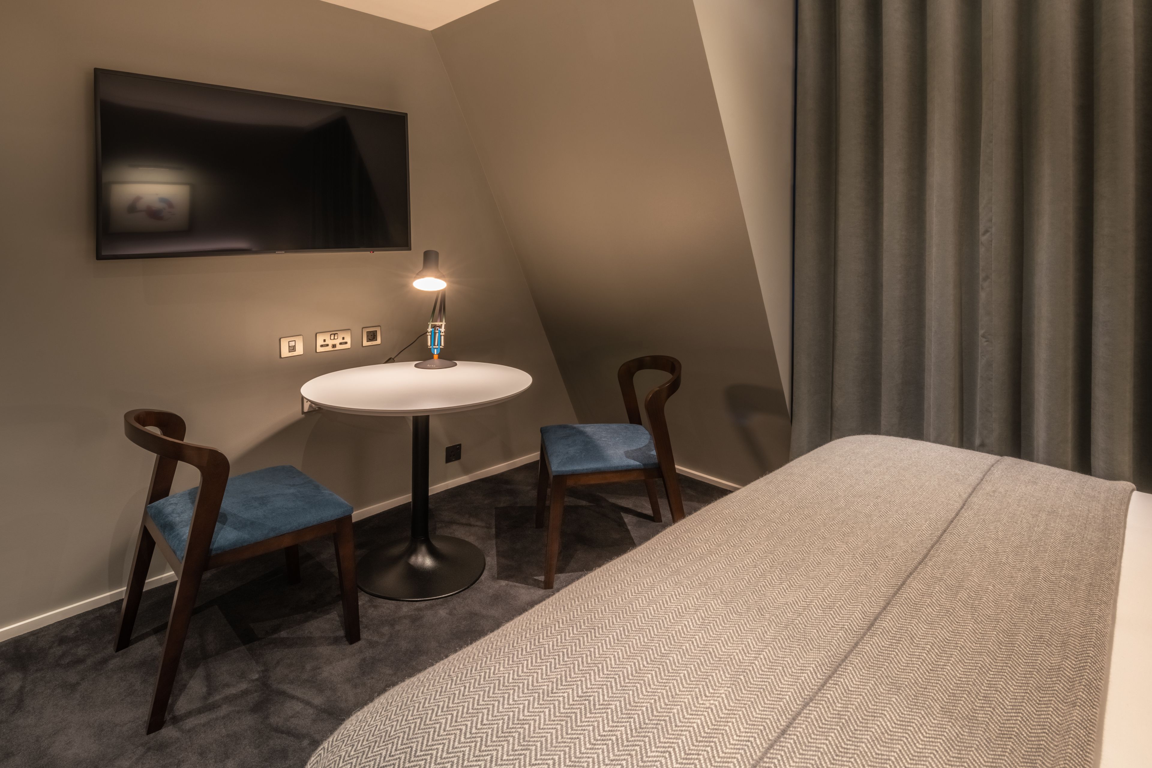 A hotel room with a bed, a round table with two chairs, a TV mounted on the wall, and a window with curtains.