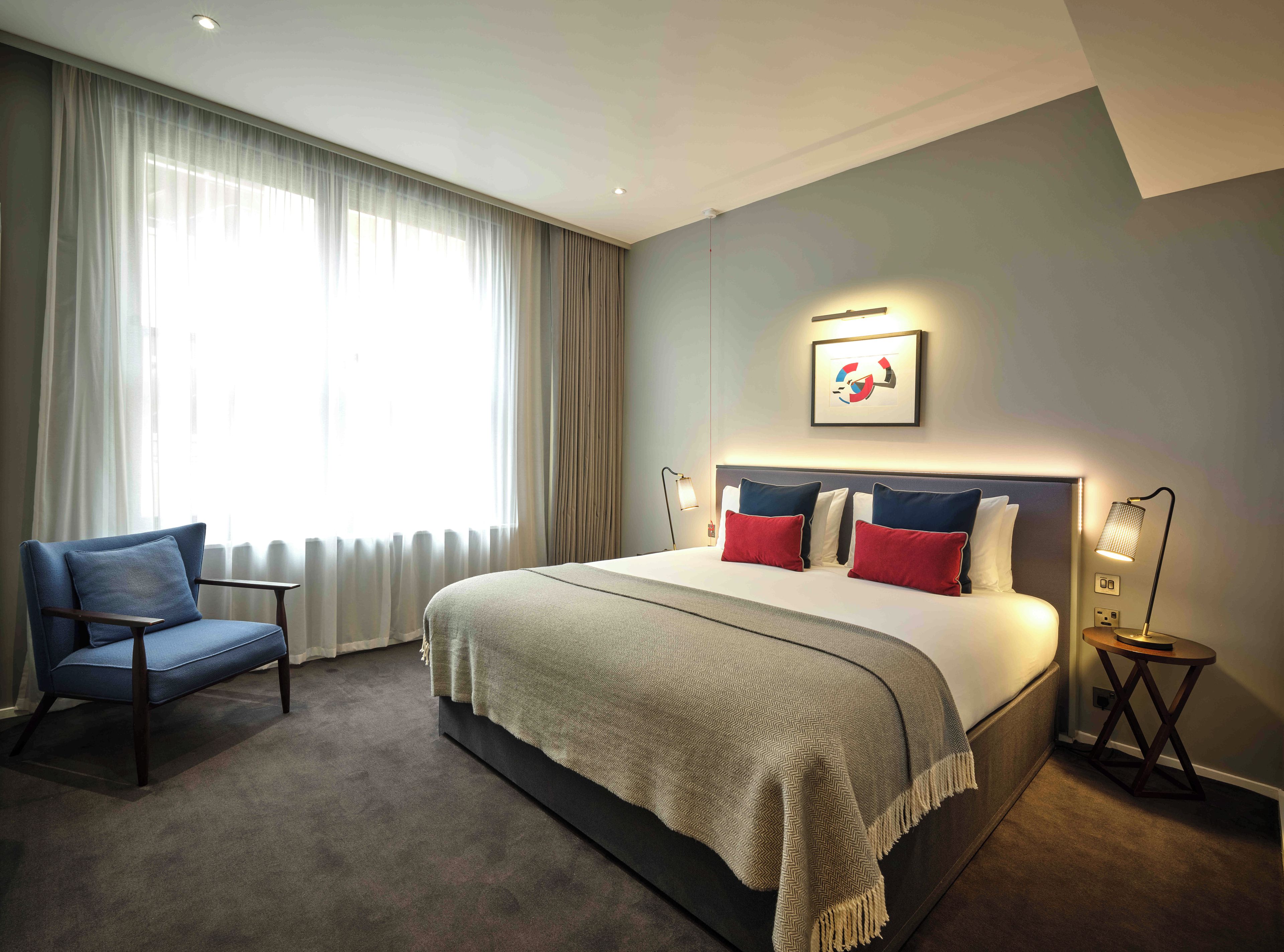 A hotel room with a large bed, a chair, a bedside table with a lamp, and a window with curtains.