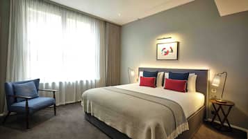 A hotel room with a large bed, a chair, a bedside table with a lamp, and a window with curtains.