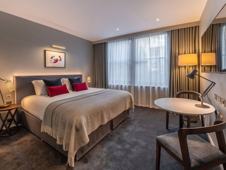 A hotel room with a large bed, a bedside table with a lamp, a round dining table with chairs, and a large window with curtains.