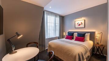 A hotel room with a bed, a desk, a chair, a lamp, a window with curtains, and a framed picture on the wall.