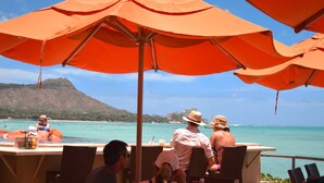 A beachside restaurant with outdoor seating and umbrellas, overlooking a clear blue sea and a mountainous backdrop. - Rare Unobstructed Diamond Head-Ocean Views, Best location, Pool, 1 minute to Beach (Honolulu)