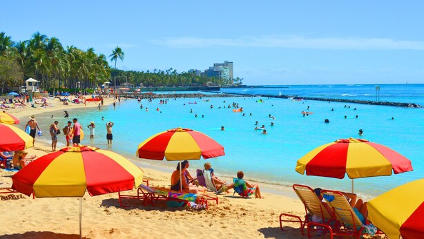 A beach with multiple red and yellow umbrellas, people swimming and sunbathing, and a clear view of the ocean and palm trees. - Rare Unobstructed Diamond Head-Ocean Views, Best location, Pool, 1 minute to Beach (Honolulu)