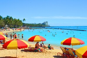 A beach with multiple red and yellow umbrellas, people swimming and sunbathing, and a clear view of the ocean and palm trees. - Rare Unobstructed Diamond Head-Ocean Views, Steps to Beach (Honolulu)