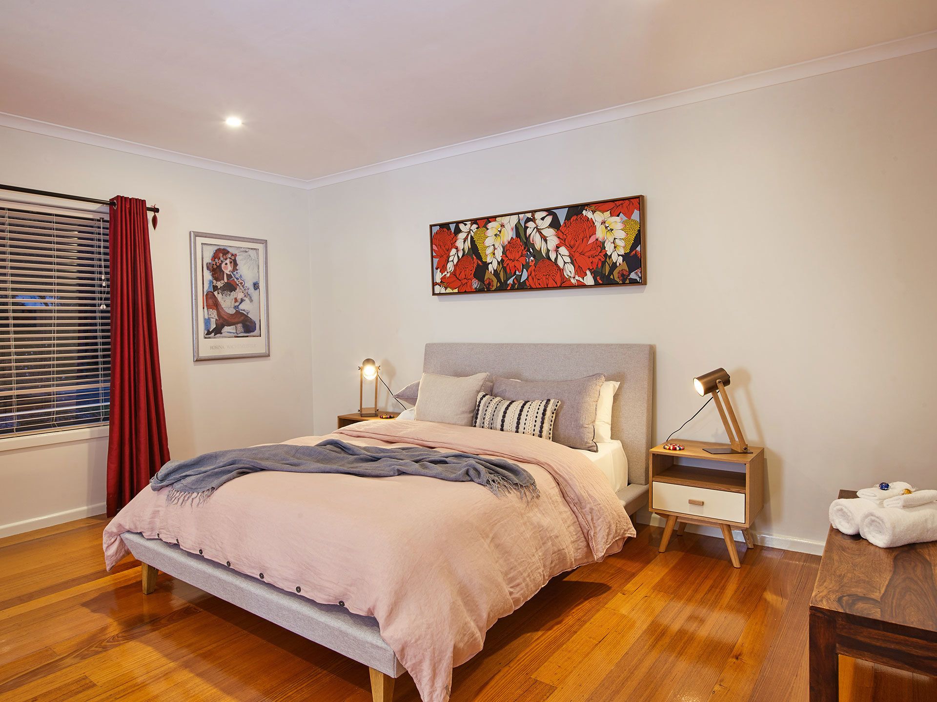 A bedroom with a bed, two bedside tables, a nightstand, a window with blinds, and a painting on the wall.