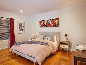A bedroom with a bed, two bedside tables, a nightstand, a window with blinds, and a painting on the wall. - Northbank House (Bright)