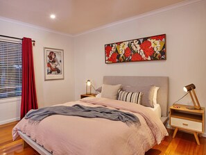 A bedroom with a bed, bedside tables, a nightstand, a window with blinds, a red curtain, and a floral painting on the wall. - Northbank House (Bright)