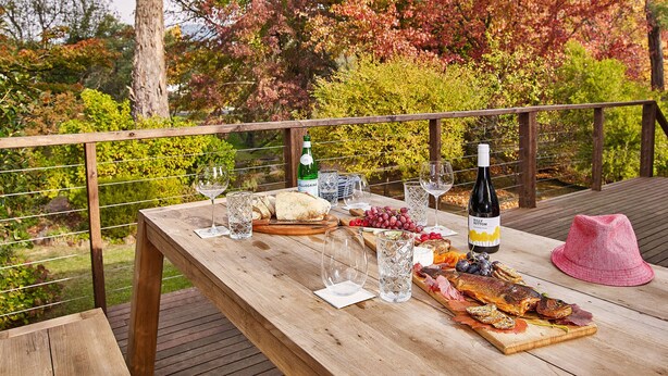 Una terraza de madera con una mesa dispuesta para una comida, que incluye una botella de vino, copas, pan y una tabla de embutidos.