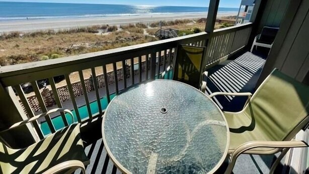Ein Balkon mit Glastisch und Stühlen, der Blick auf einen Strand und das Meer freigibt.