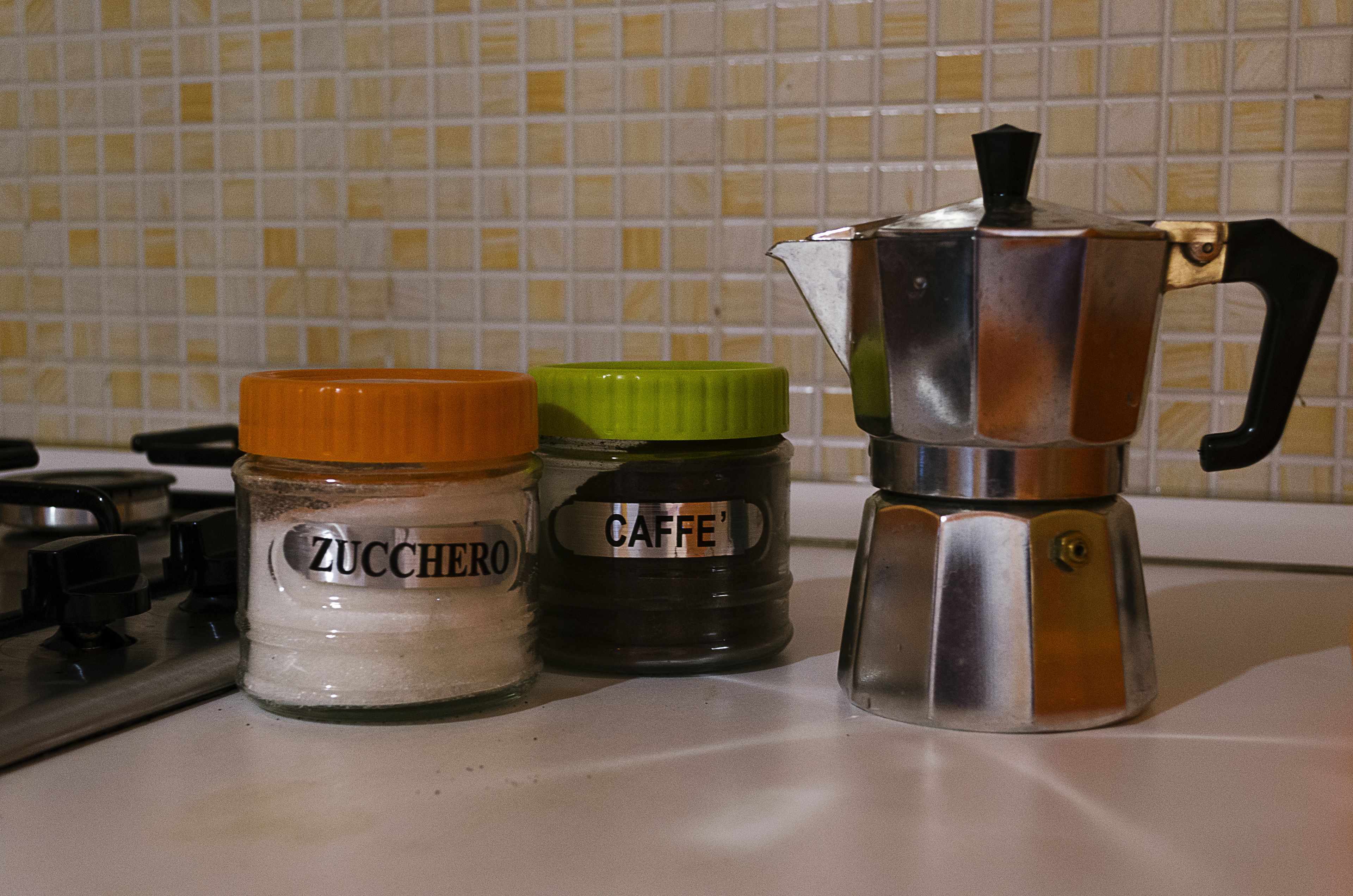 A kitchen counter with a stove, two jars labeled 'ZUCCHERO' and 'CAFFE'', and a metallic coffee pot.
