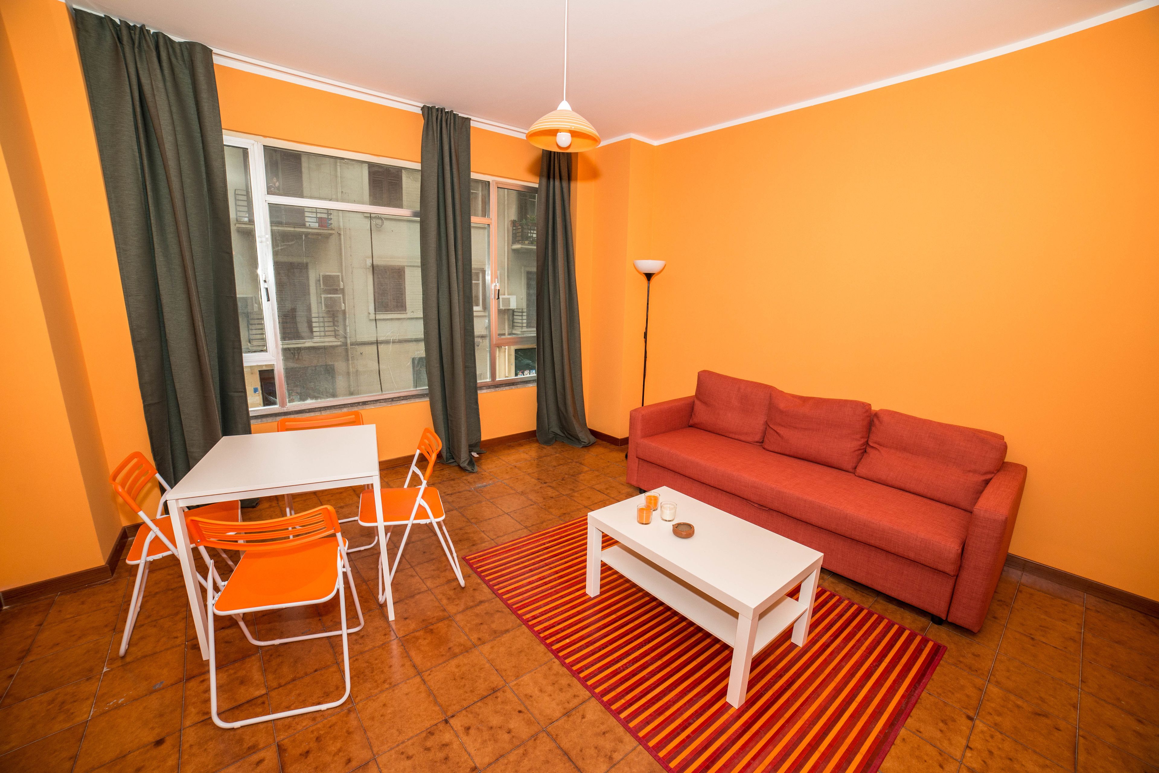 A small living room with a red sofa, white coffee table, orange chairs, and a window with curtains.