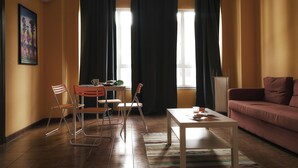 Ein Wohnzimmer mit einer Couch, einem Tisch und zwei Stühlen. Es gibt ein Fenster mit Vorhängen, ein Bild an der Wand und eine Klimaanlage.
