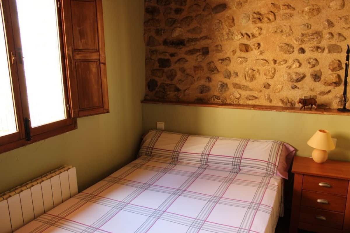 A bedroom with a bed, a wooden nightstand, a lamp, a stone wall, and a window with wooden shutters.