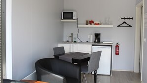 A compact kitchen with a microwave, coffee maker, and a small shelf with various items. There is a dining table with chairs and a fire extinguisher mounted on the wall. - Systrakot (Talknafjordur)